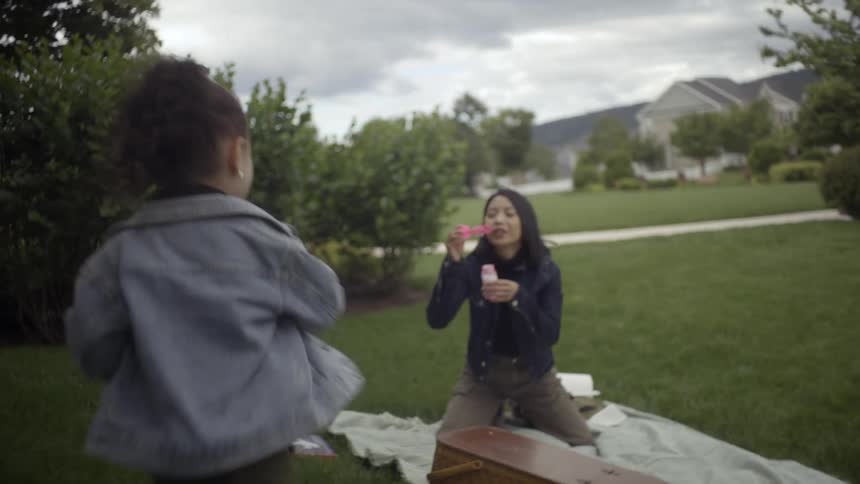 A mother blows bubbles while her joyful daughter runs around playfully during a picnic at the park