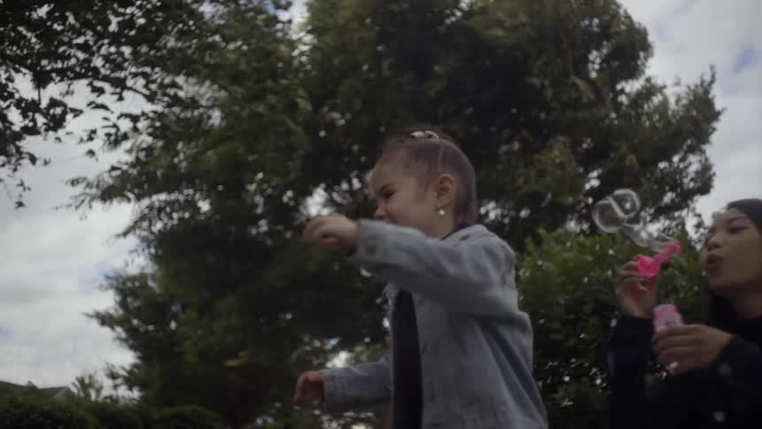 Mother blows bubbles as her daughter joyfully runs laughs and plays in a sunny park