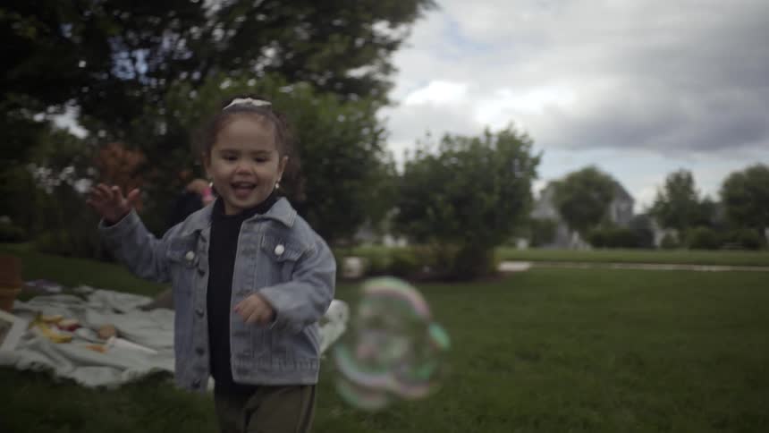 A young child chases bubbles blown by an adult in a grassy park