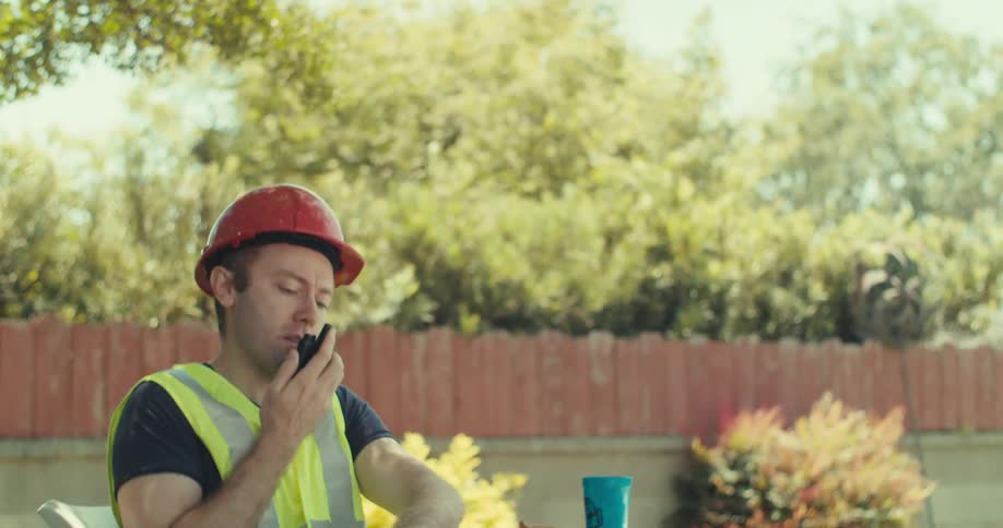 A construction worker wearing safety gear sits down at a table as he talks on a radio and starts to write with a pen