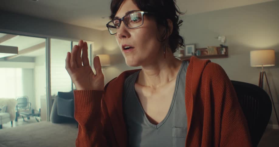 A young woman wearing glasses sitting in front of a computer looking surprised in her sitting room at midday