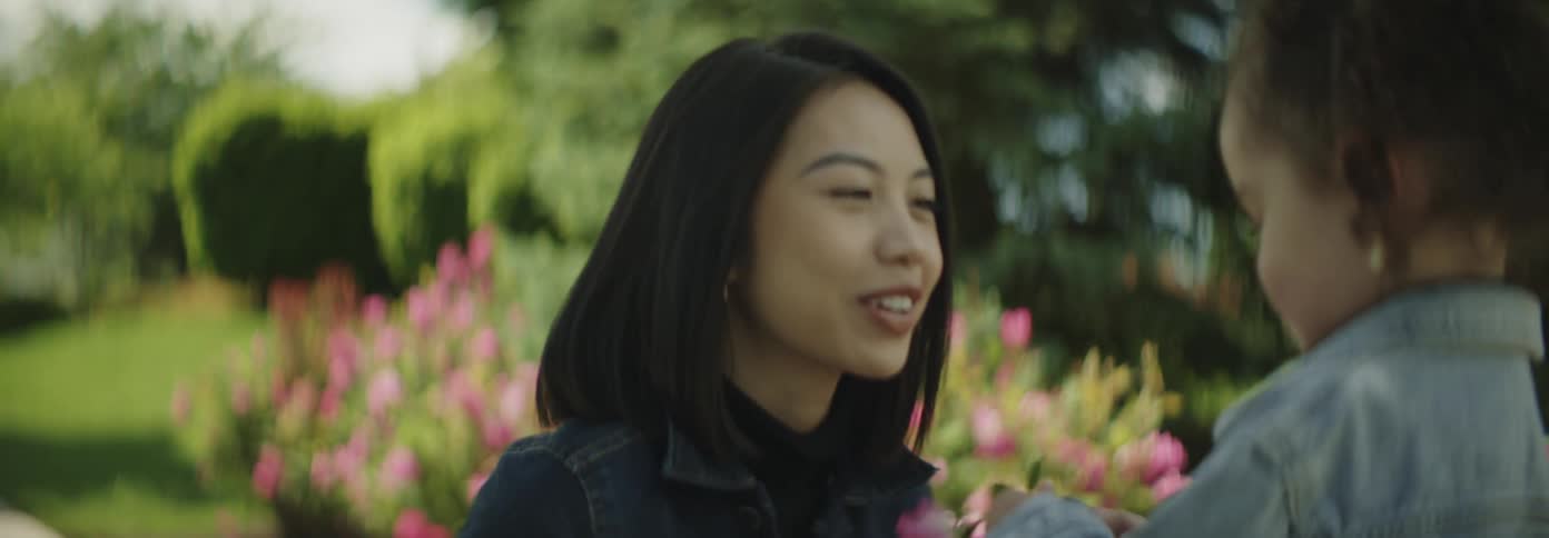 An asian young woman squats on the sidewalk at midday talking to her child while holding a flower with a flower garden lining the side of the road
