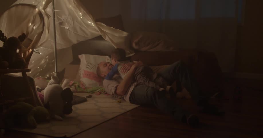 Man holding an infant while sitting on the floor and then lying back to rest while talking to the child