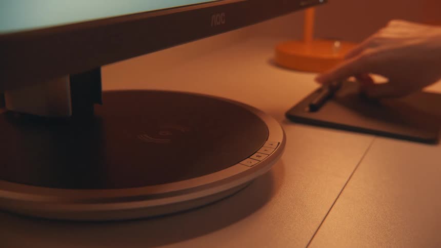 A person uses a stylus on a digital tablet beside a monitor in a warmly lit workspace