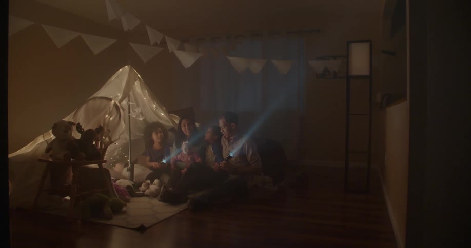 A tent is set in this dark room and a family is sitting on the floor