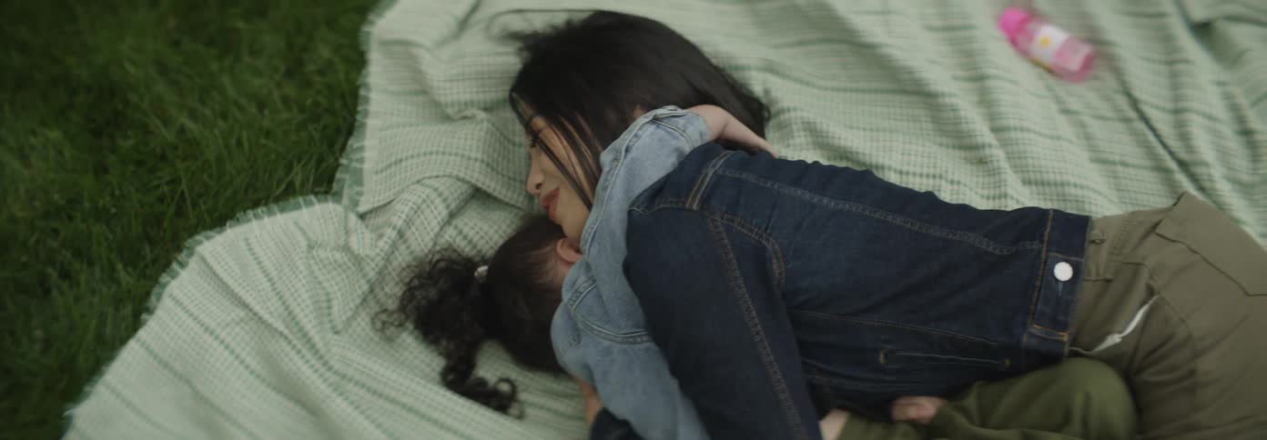 A young girl and an adult woman are lying on a green patterned blanket smiling and laughing together as the girl embraces the woman