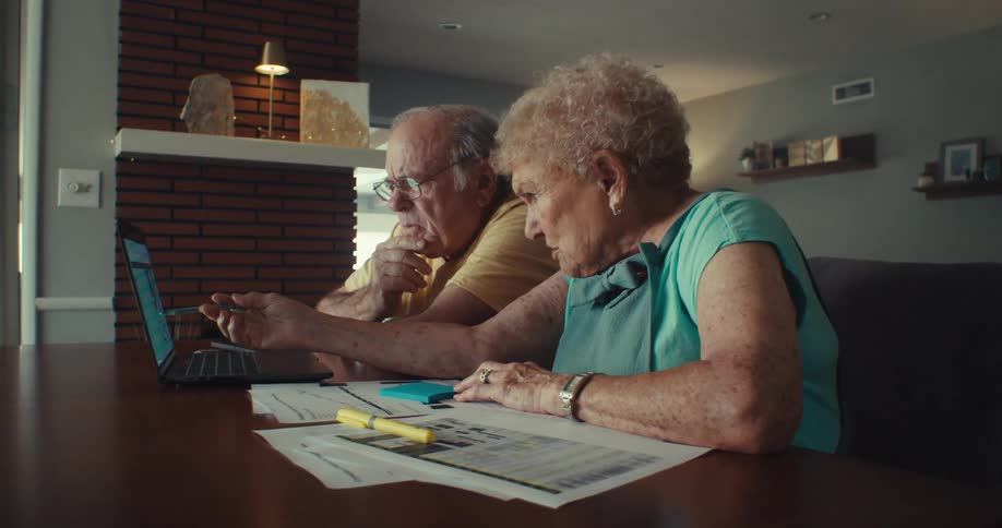 Elderly couple looks at a table displayed on a laptop while working on paperwork at their dining table during the daytime
