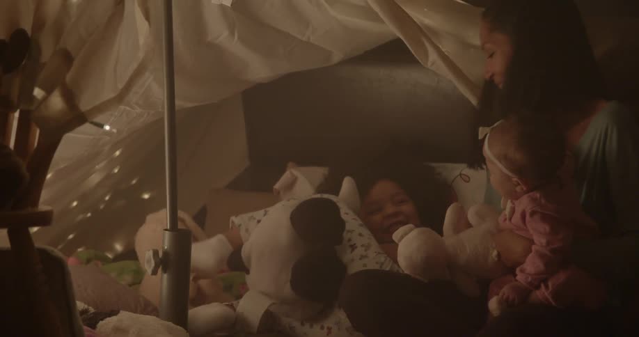 Woman playing with young children that have stuffed animals as they are all indoors under a homemade tent