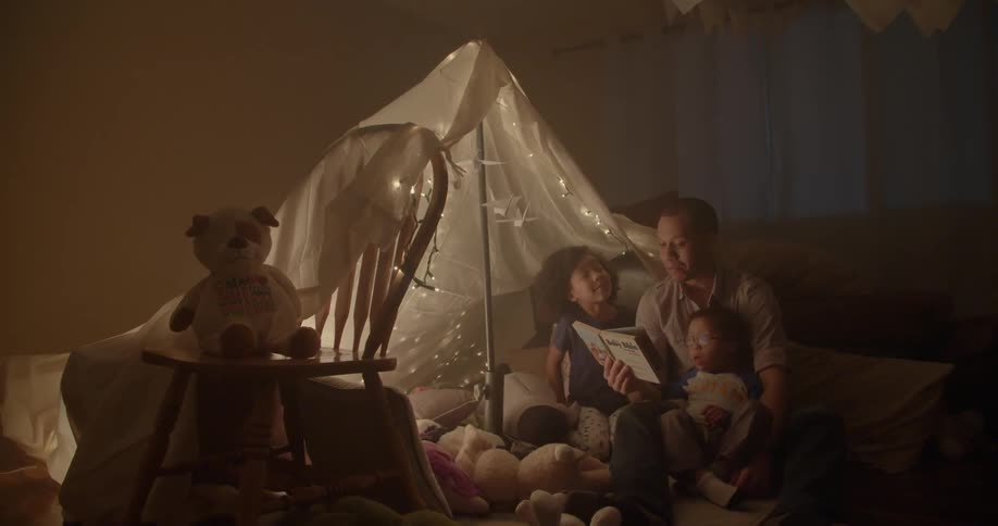 A father and children read a book under a blanket tent