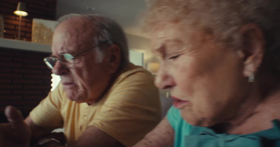 An older couple sits together in a cozy living room looking worried as they have a serious conversation