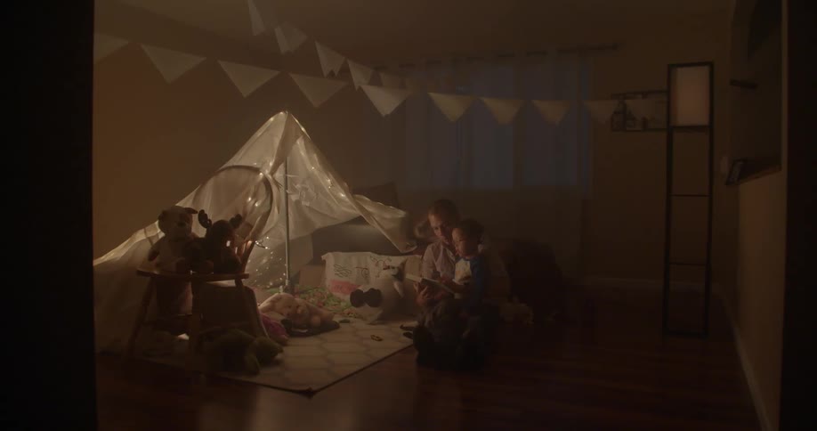 A man and child sit on the floor near a blanket tent and read a book