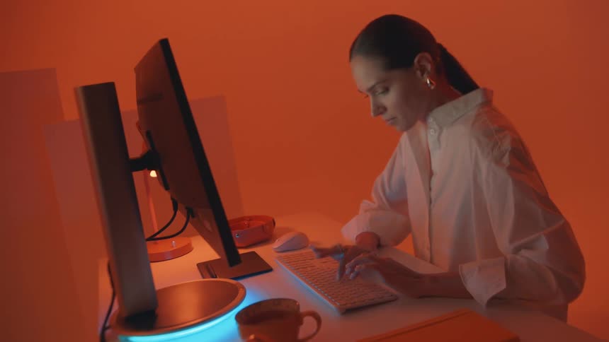 A woman works on a computer in a warmly lit orangehued room deeply focused on typing