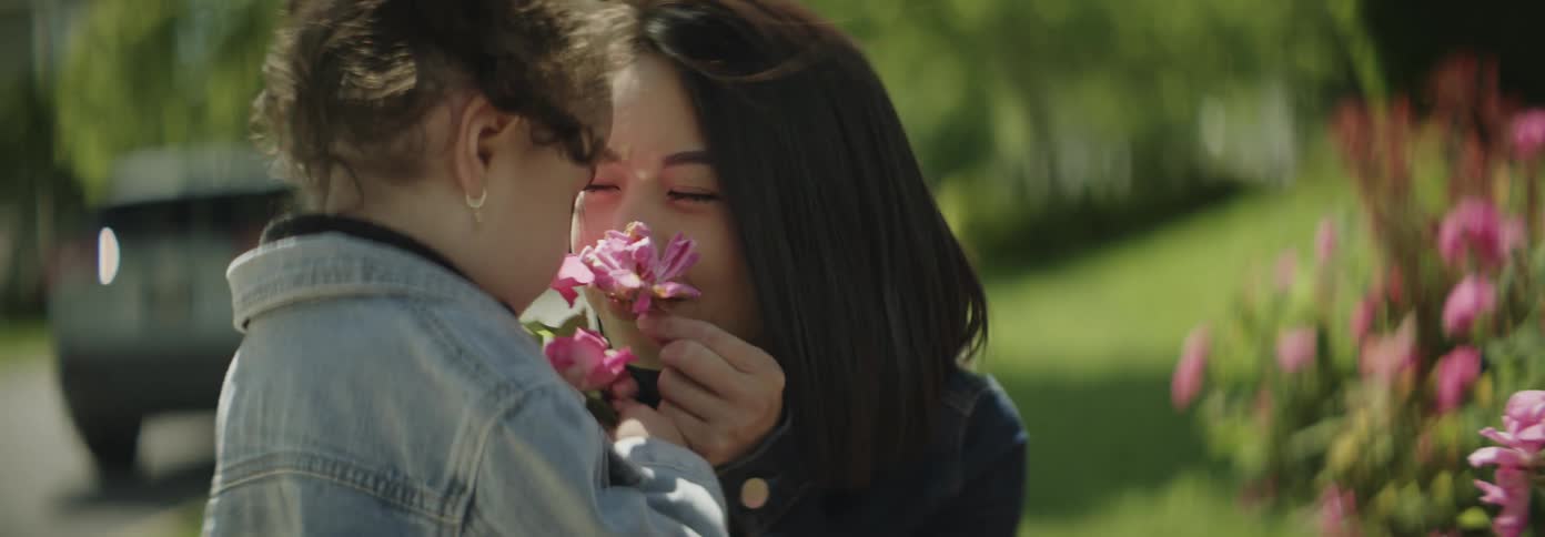 A young woman is showing pink flowers to a toddler girl on a suburb sidewalk