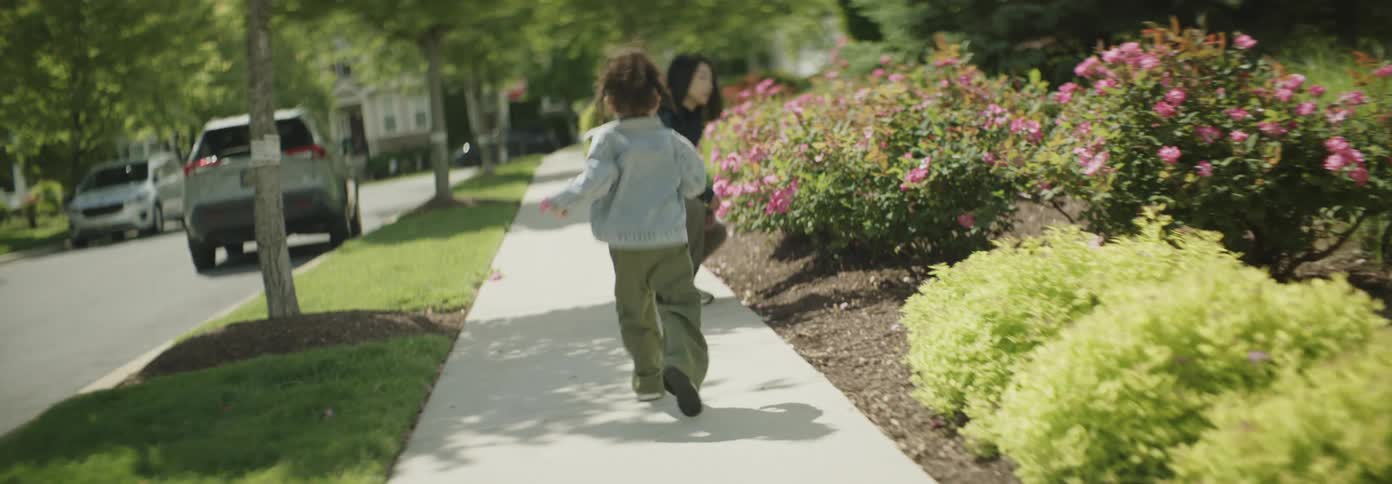 A girl runs up to her mom to smell flowers in a bush