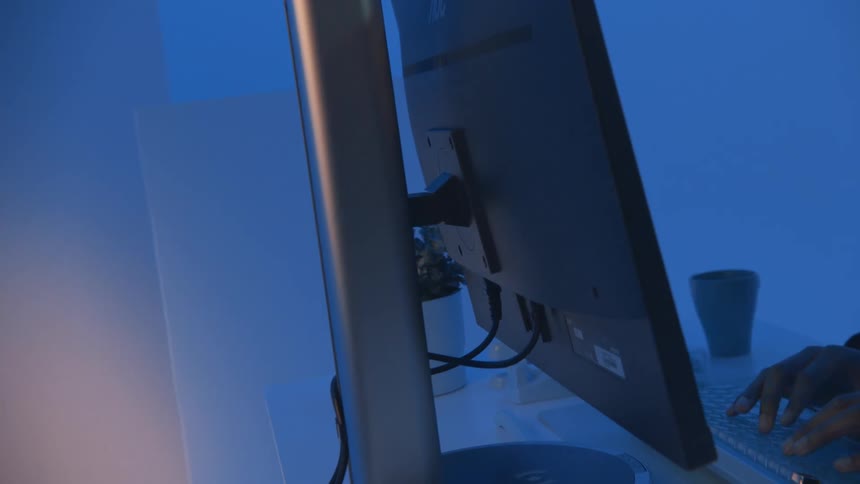 A computer monitor is seen from behind with a persons hands typing on a keyboard in a bluelit environment before turning around and revealing a website