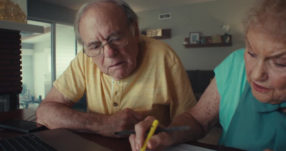 An older woman and man sitting at a table the woman is using a highlighter on a sheet of paper