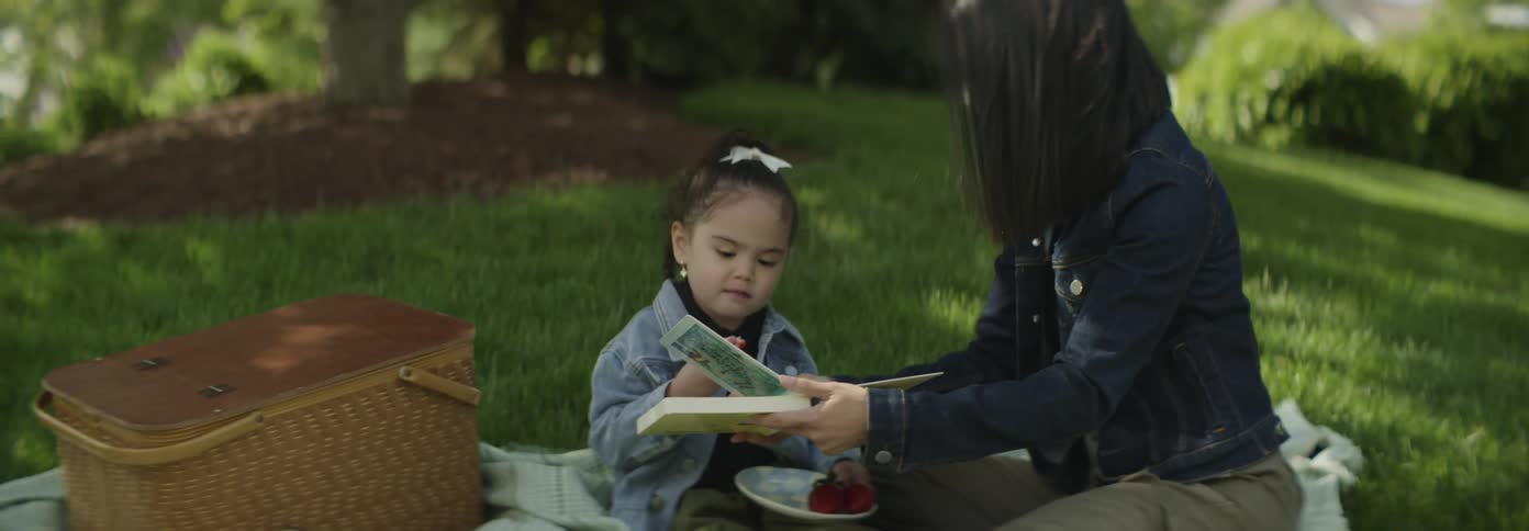 Mother reads a book to her daughter during a joyful picnic outdoors while the child enjoys food