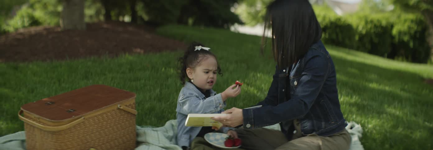A woman and a young child sit on a blanket in a grassy area looking at a book and eating strawberries near a picnic basket