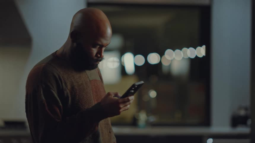 A bald man with a beard and mustache looks at a smartphone in a dimly lit indoor setting