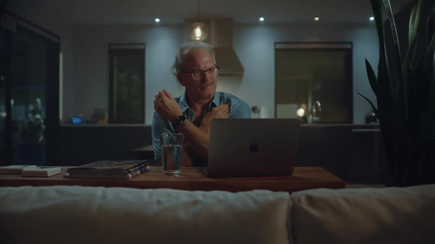 A man is in pain and is rubbing his shoulder in front of the laptop