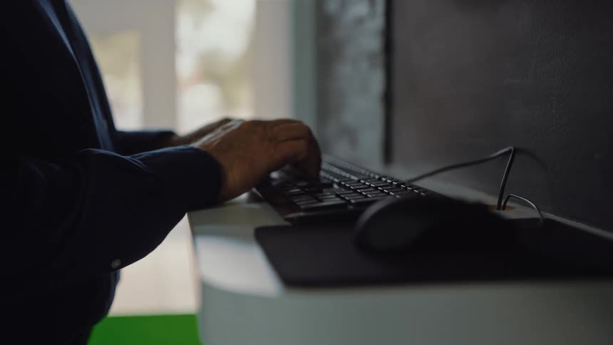 Hands type on a black computer keyboard situated on a white desk with a mouse and mousepad