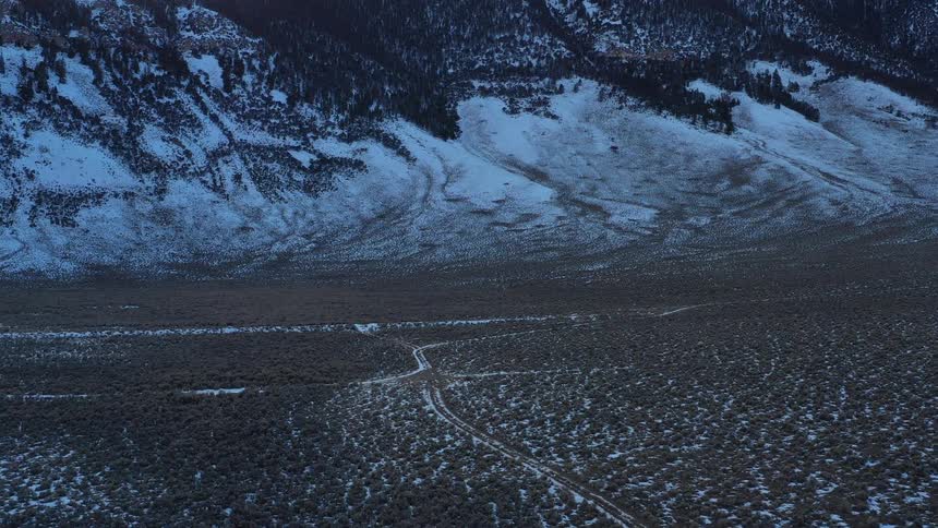 An aerial view reveals a snow dusted desolate landscape featuring a winding dirt path that extends towards a dark partially snow covered mountain range in the distance