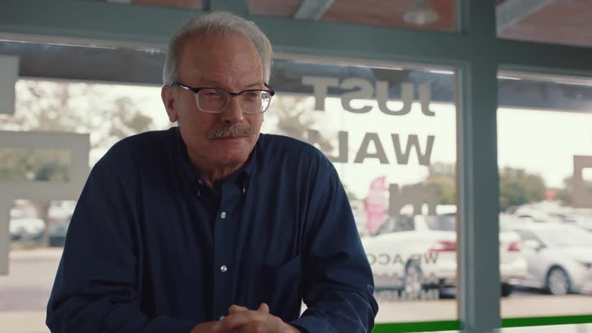 A smiling older man in glasses and a blue shirt is talking to someone inside a storefront