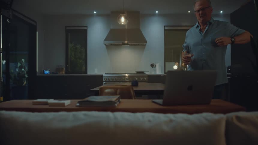 An elderly man works intently on a laptop at a dining table in a dimly lit modern kitchen at night