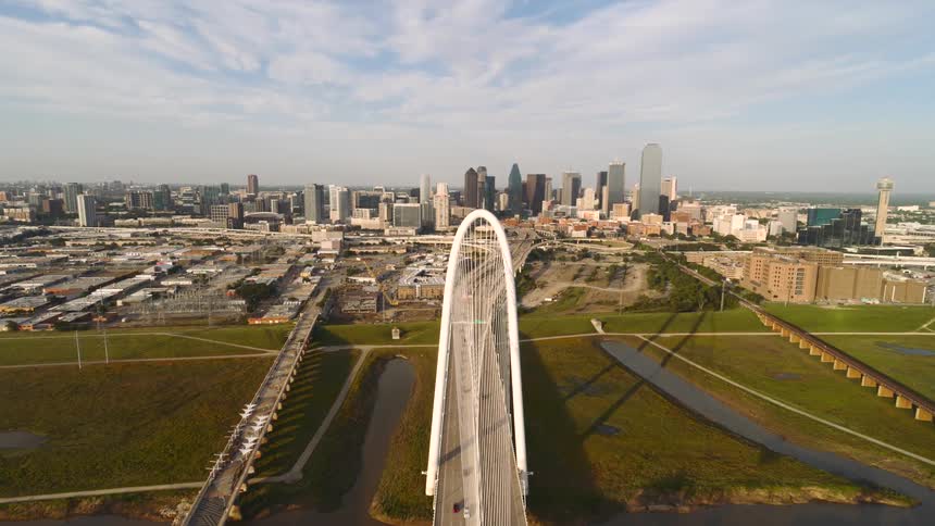 An aerial view captures the dallas skyline and the margaret hunt hill bridge featuring traffic and a waving texas flag set against green fields and a river under a clear sky