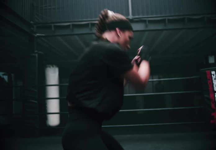 A woman with braided hair and a black headband shadow boxes in a dark industrial style boxing ring moving swiftly as she practices her punches
