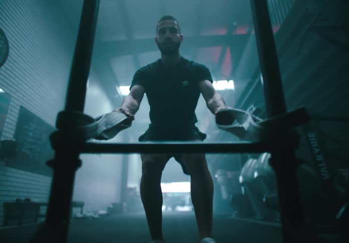A man intensely pushes a piece of gym equipment towards the camera in a dimly lit hazy fitness facility viewed from the perspective of the equipment itself