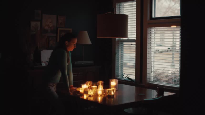 A woman leans over a table filled with lit candles in a dimly lit room staring thoughtfully