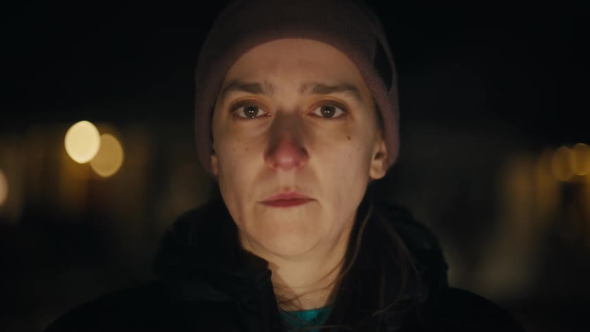 A person wearing a dark beanie and jacket looks directly at the camera with a concerned expression in a dark outdoor setting with blurred lights in the background
