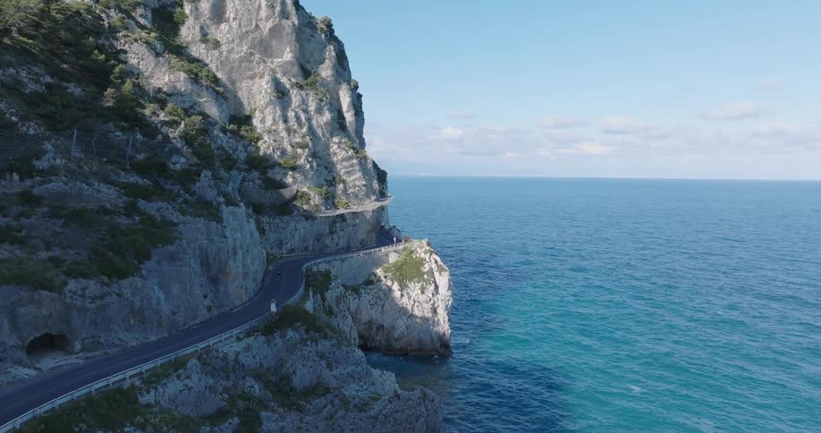 An aerial shot follows a winding coastal road carved into a steep cliffside with vehicles navigating its curves alongside the expansive blue ocean