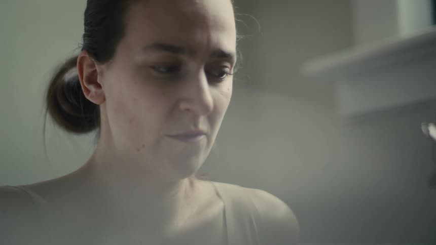 A closeup shot shows a womans face looking downwards with her hair pulled back and a softly focused dimly lit background