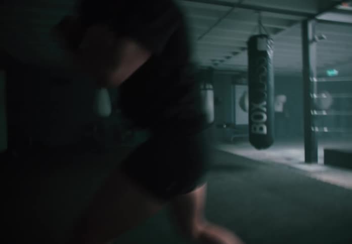 A boxer in a dimly lit gym shadow boxing near a boxing ring and hanging punching bags