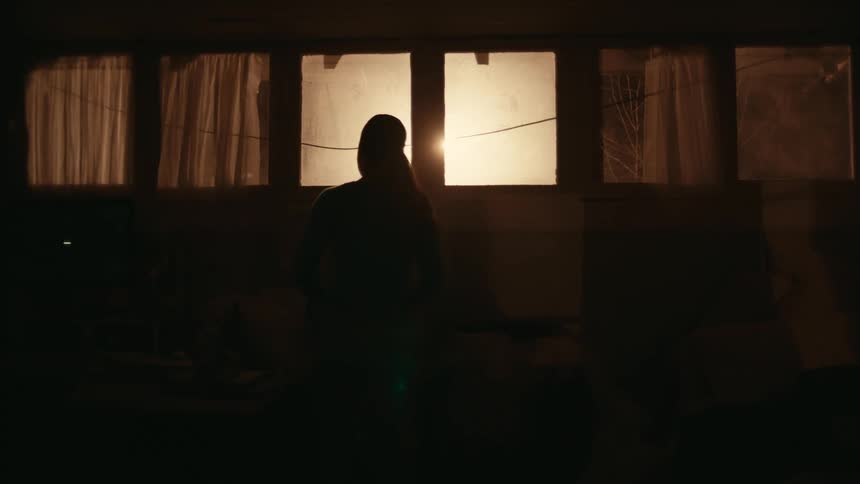 A person stands silhouetted in darkness in front of brightly lit windows