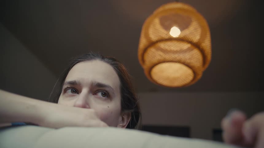 A young woman sits on a couch in the living room with a ceiling lamp illuminating the room