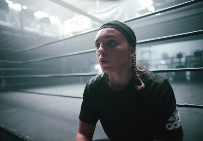 A woman sits exhausted on the edge of a boxing ring in a dimly lit gym