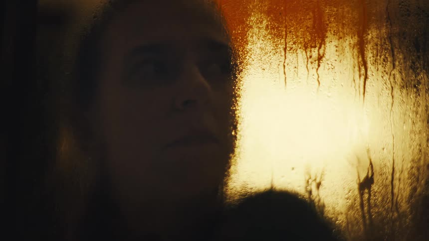 A persons face is seen through a wet glass surface illuminated by a warm fiery light
