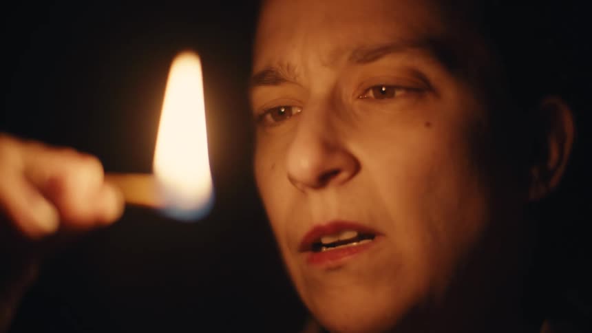 A woman holds a lit match in a dark room staring at it for a moment before blowing it out leaving the room in darkness