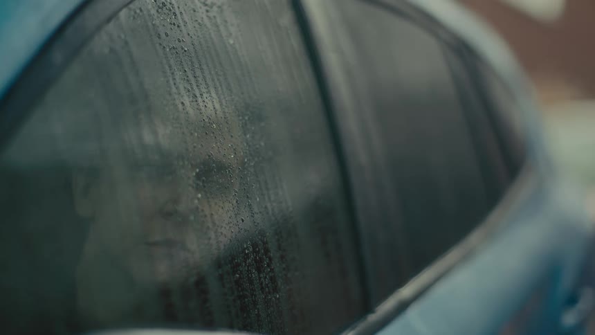 A young man sits on the drivers side of a parked car as rainwater drips down the window glass