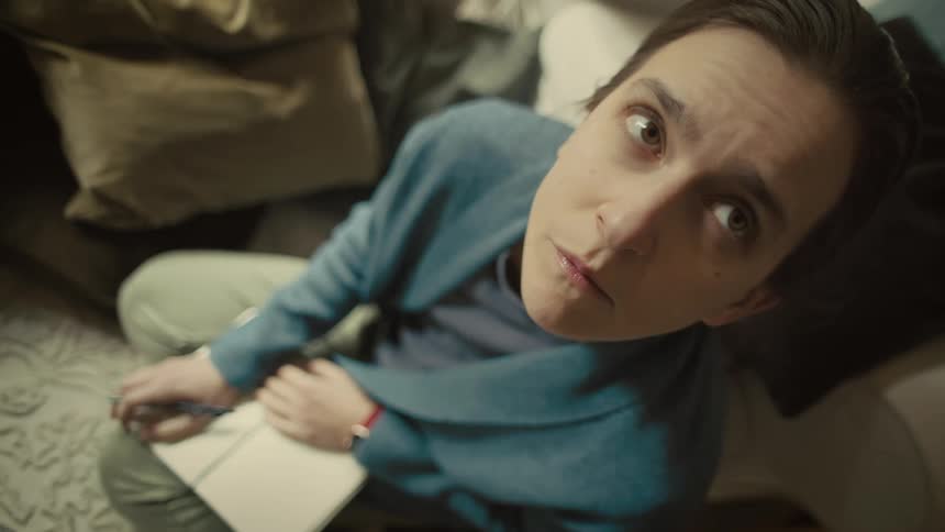 A woman sits indoors looking up thoughtfully while holding a notebook