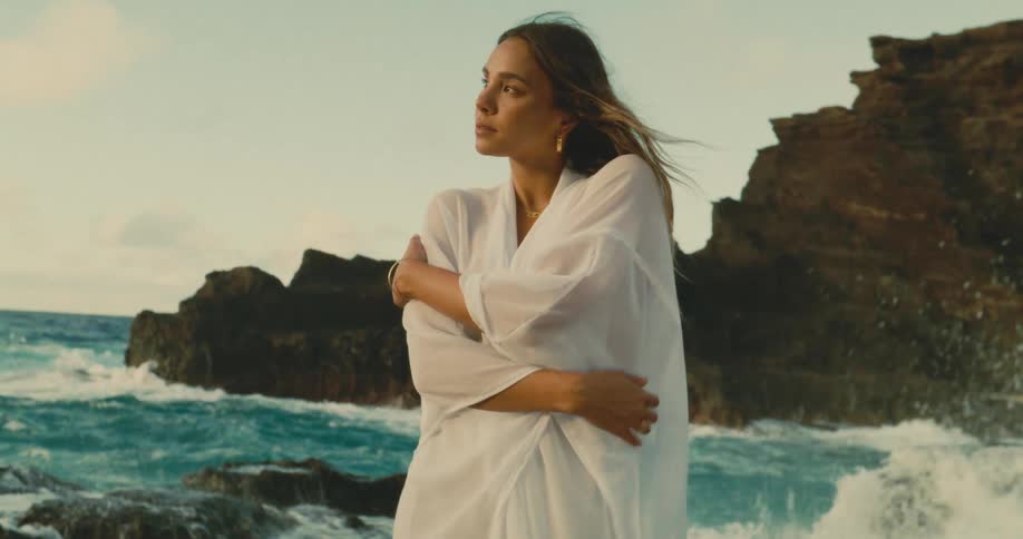 A serene woman in a flowing white robe stands with her eyes closed embracing herself on a rocky shoreline as waves crash in the background under a soft golden sky