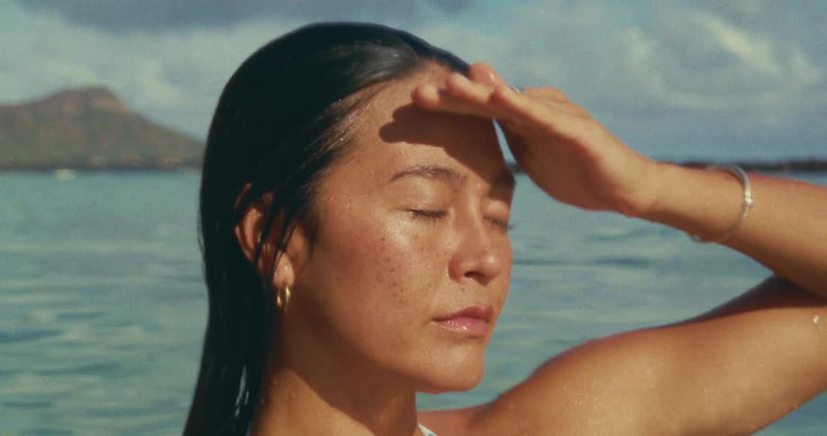 A woman stands in shallow ocean water shielding her eyes from the sun