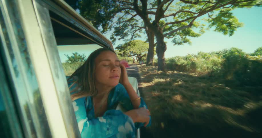 Female passenger holding a flower leans out the window of a car during a road trip through the rural countryside on a sunny day