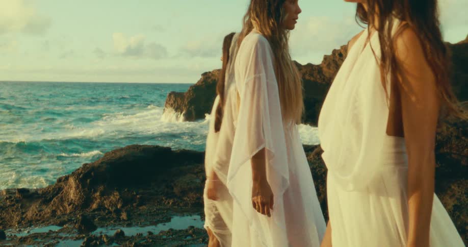Three women in white sheer dresses walk along a rocky shoreline at sunset