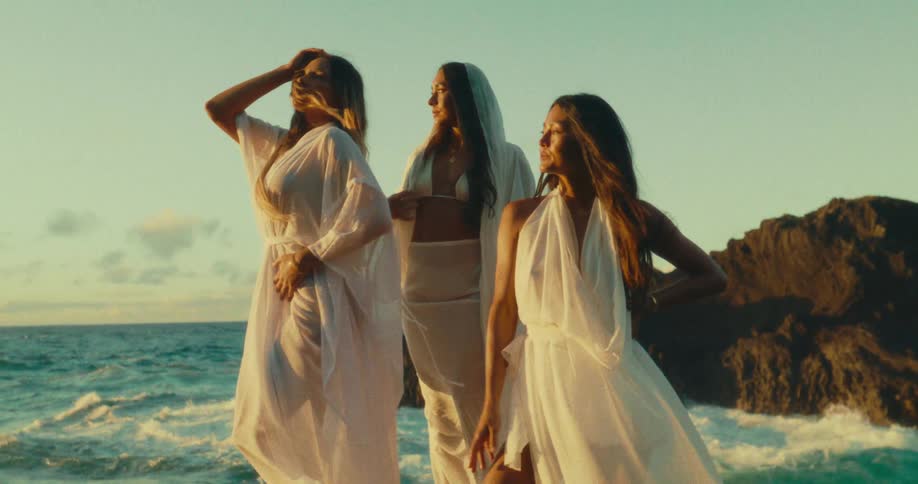 Three women in flowing white garments pose confidently by crashing waves on the coast