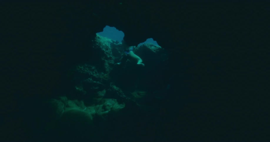 A person swims upwards through an opening in a dark underwater cave silhouetted against the brighter water above