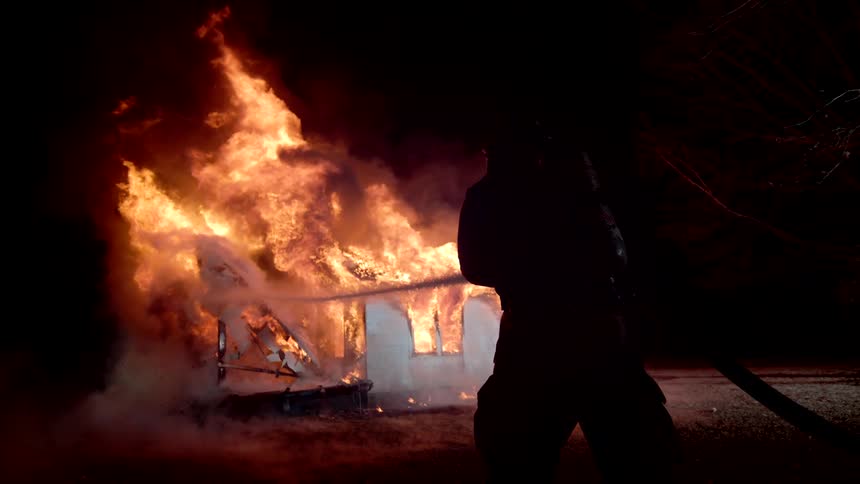 At night firefighters battle a large house fire with one spraying water directly onto the blazing structure as intense flames and smoke engulf the building